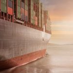 A colossal cargo ship loaded with containers navigates through calm waters against a vibrant sunset sky.